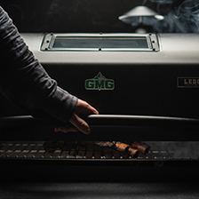 Person opening Green Mountain Grills pellet grill, smoke rising, showcasing grilling experience and quality craftsmanship.